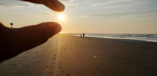 Optical illusion of hand holding sun over sea against sky during sunset