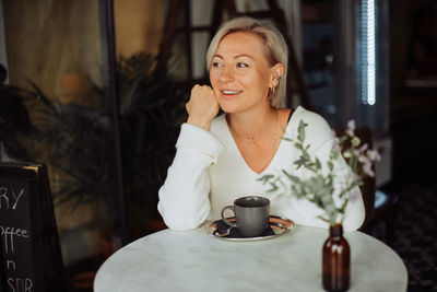 Blond woman smiling while sitting at table in cafe with cup of coffee