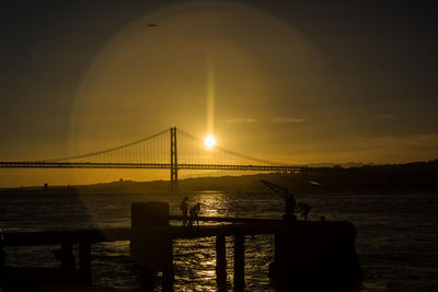 Silhouette suspension bridge over sea against sky during sunset