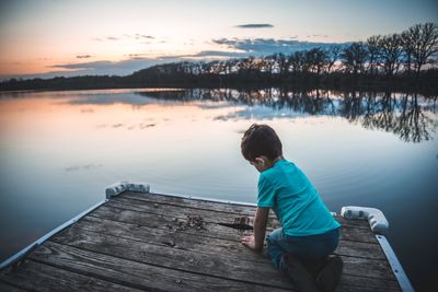 Boy on jetty over lake against sky during sunset