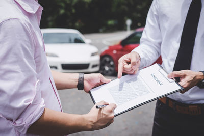 Midsection of man signing insurance paper held by agent at parking lot