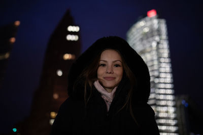 Portrait of young woman against illuminated buildings in city at night