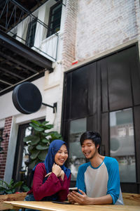 Low angle view of smiling couple using smart phone while sitting at sidewalk cafe