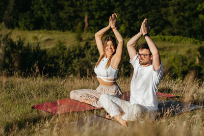 Cheerful woman and happy man raising hands up over heads, young adult couple meditating outdoors