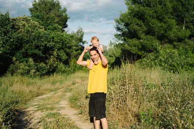 Content young man standing with little child on shoulders on sandy road in village on sunny day and looking at camera