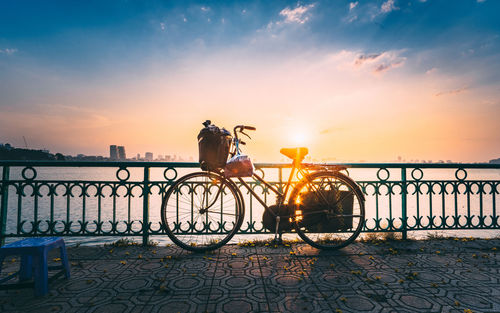 Bicycle by railing by water during sunset