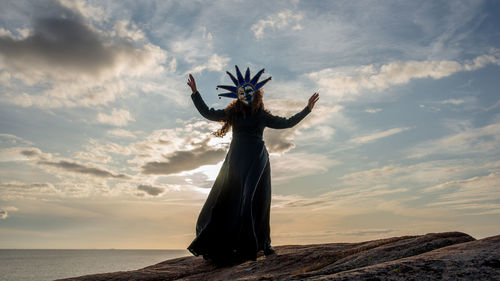 Woman wearing mask standing at beach against sky during sunset