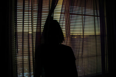 Rear view of silhouette woman looking through window at home