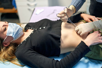 High angle of anonymous body modification artist pinching belly button of female client with face mask during piercing procedure in salon