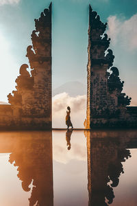 Reflection of woman amidst built structure on lake against sky