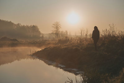 Rear view of silhouette man standing by lake against sky at sunrise