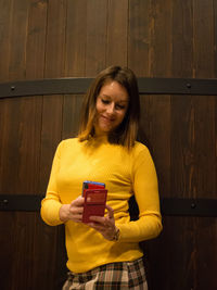 Young woman using phone while standing against wall