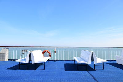 Empty chairs and tables by sea against blue sky