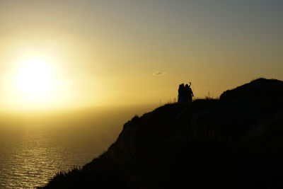 Silhouette people on rock by sea against sky during sunset