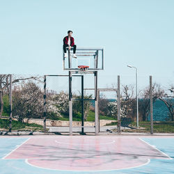 Man sitting on basketball hoop against sky