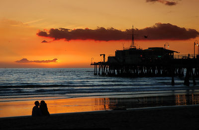 Silhouette couple sitting at beach by santa monica pier against sky during sunset