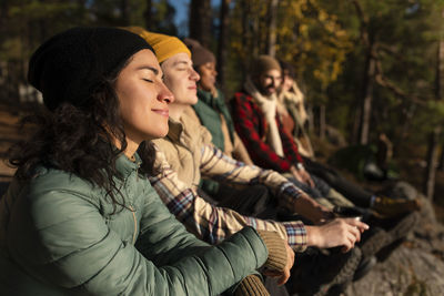 Side view of smiling woman with eyes closed enjoying sunlight while sitting by friends in forest