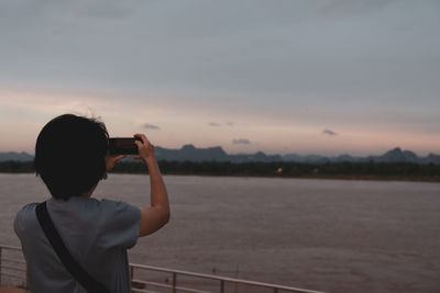 Rear view of man photographing sea against sky during sunset