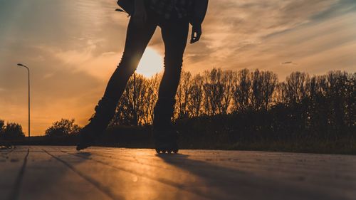 Low section of person skating by trees against sky during sunset