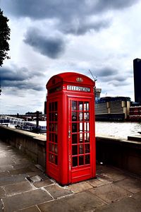 Red telephone booth by river against cloudy sky