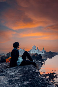 Man sitting on rock against sky during sunset