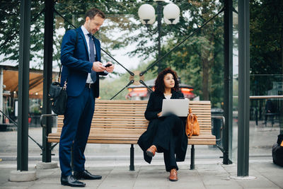 Full length of business people using smart phones while waiting at bus stop