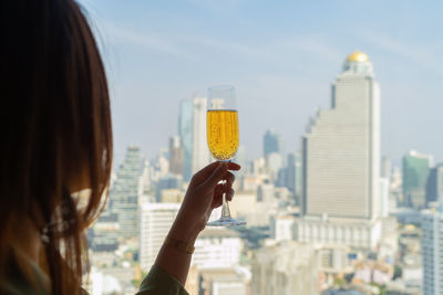 Rear view of woman holding glass against buildings in city