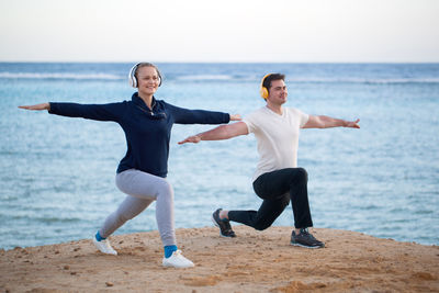 Couple listening music while exercising at beach against sky during sunset