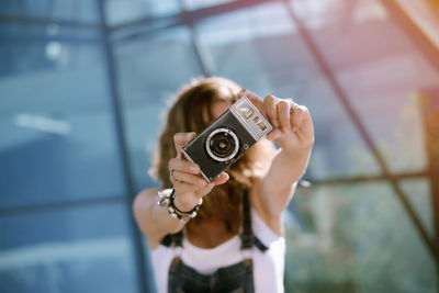 From above of young unrecognizable female holding photo camera in outstretched hands and taking picture while standing against glass wall