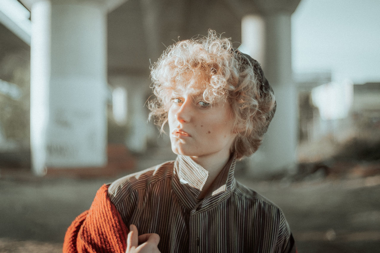 headshot, one person, focus on foreground, portrait, lifestyles, real people, leisure activity, day, hairstyle, architecture, built structure, hair, front view, casual clothing, outdoors, young adult, building exterior, blond hair