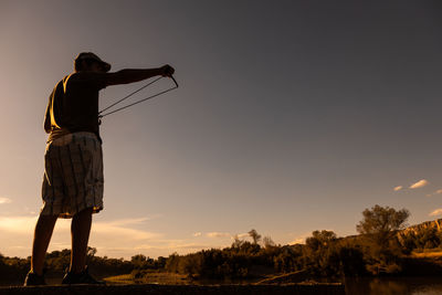 Rear view of man standing on field holding slingshot against sky during sunset