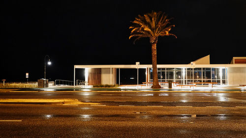 Illuminated street by building against sky at night