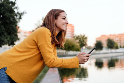 Smiling caucasian woman wearing yellow pullover using mobile phone device outdoors in park 