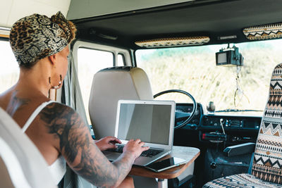 Back view of optimistic female in white dress typing on modern netbook while sitting near window in light camping van during road trip