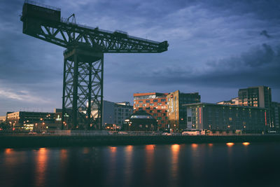 Illuminated buildings by river against sky at dusk