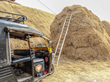 A black rickshaw is parked in front of a big pile of straw, in front of which is a wooden ladder.