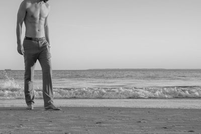 Low section of shirtless man standing at beach against sky