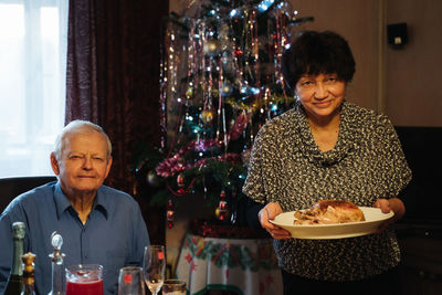 Positive senior woman holding plate with roasted chicken and smiling husband sitting at table during family dinner at home with christmas tree in background
