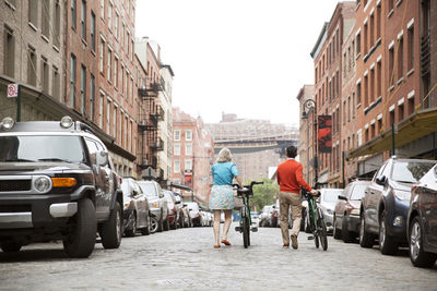 Rear view of mature couple walking with bicycles on city street