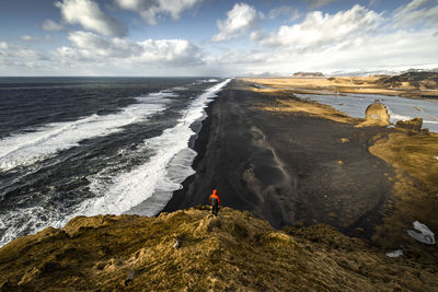 From above unrecognizable traveler in warm clothes standing on rocky cliff on black sandy beach and enjoying spectacular ocean view during vacation in iceland person