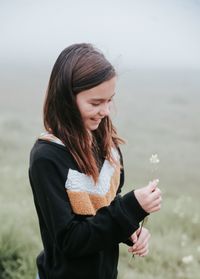 Smiling girl holding flower while standing on field during foggy weather