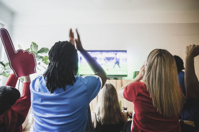 Rear view of excited soccer fans watching match with friends at home