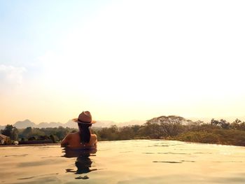 Rear view of woman looking at landscape while resting in infinity pool against sky during sunset