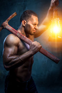 Shirtless muscular worker holding lantern and pick axe while standing against wall