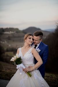 Bride holding flower bouquet with groom embracing against sky