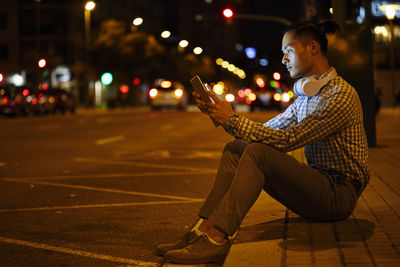 Young man sitting at roadside in the city at night using tablet