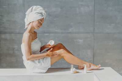 Full length of woman wearing towel applying moisturizer in bathroom