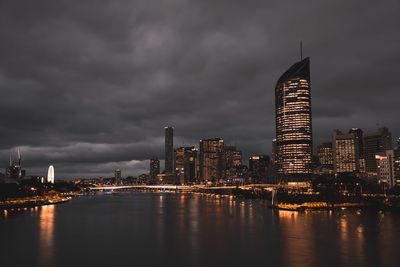 Illuminated city at waterfront against cloudy sky