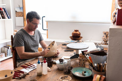 Man sitting at table with brushes and drawing sketches on handmade ceramic plate