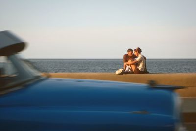 Young couple sitting on retaining wall by sea against sky